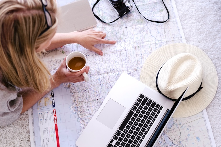 Woman looking at a map with coffee, a hat, and a laptop - one of the ways to plan for a European road trip.