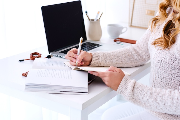 Woman writing in a notebook, next to a laptop, phone, calculator, notebooks, and stationery - things you need when using a paper planner and Google Calendar together.