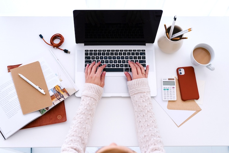 Woman typing on a laptop, next to a phone, calculator, notebooks, and stationery - things you need when using a paper planner and Google Calendar together.