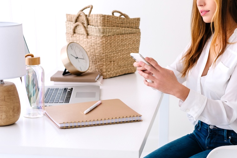 Woman holding a phone with laptop, clock, notebook, pen and baskets in front of her - things you need to prepare for back to school.