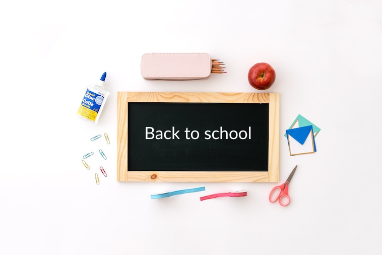 Blackboard saying back to school surrounded by glue, pencils, apple, envelopes, scissors, ribbon, paper clips - school supplies you can buy when making school traditions.