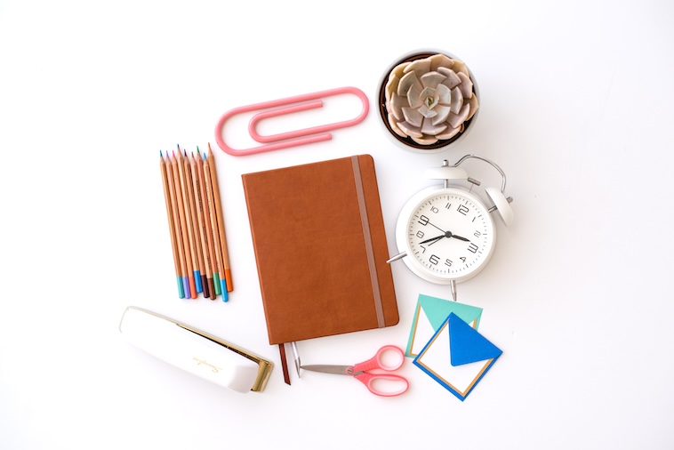 Notebook surrounded by plant, clock, envelopes, scissors, stapler, pencils - school supplies you can buy when making school traditions.