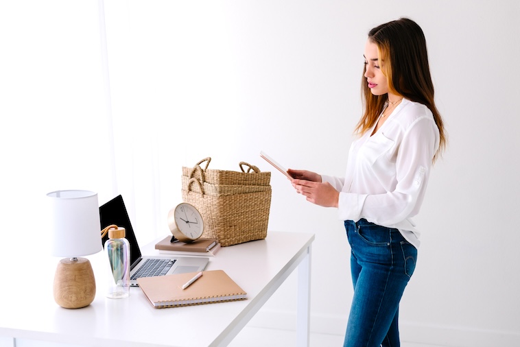 Desk with lamp, laptop, water bottle, clock, notebook, baskets and woman holding tablet - things you need when using time blocking.