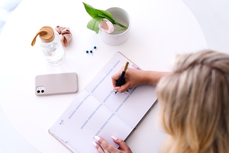 Woman writing in notepad with phone, bottle of water pink flower - things you need when writing goal ideas.