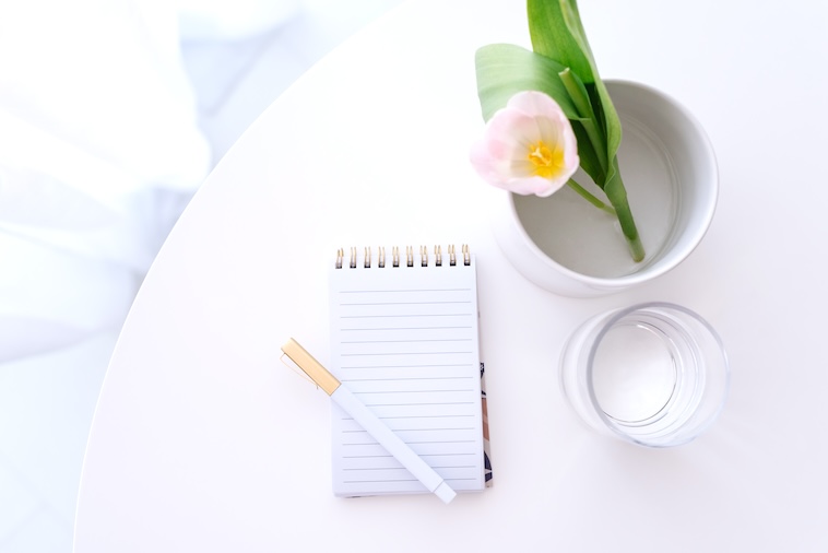 Table with notepad, pen, glass of water, pink flower - things you need when writing goal ideas.