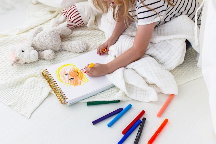 Young girl drawing with markers, a soft toy and sitting on a blanket - one of the things kids can do at home.