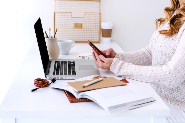 Woman holding a phone, next to a laptop, notebooks, and stationery - things you need when using a paper planner and Google Calendar together.