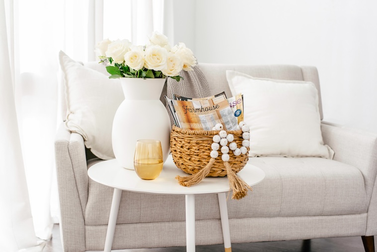 Grey sofa, coffee table with vase and white roses, glass, and wicker basket holding magazines - things you can remove when you want to simplify your home.