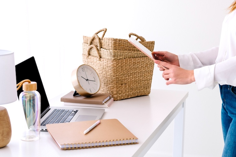 Desk with lamp, laptop, water bottle, clock, notebook, baskets and woman holding tablet - things you need when using time blocking.